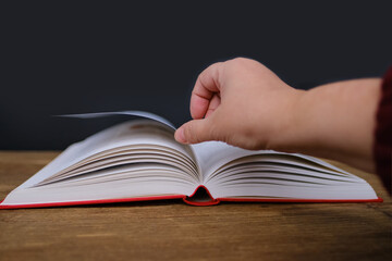 open paper book with red cover lies on a wooden table closeup, hand flipping pages, reading concept, copy space