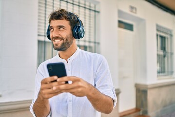 Handsome man with beard wearing casual white shirt on a sunny day smiling happy outdoors using smartphone listening to music wearing headphones