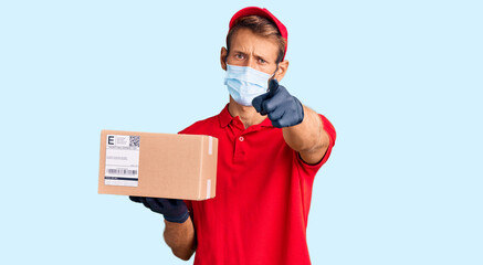 Handsome blond man with beard holding delivery box wearing medical mask pointing with finger to the camera and to you, confident gesture looking serious
