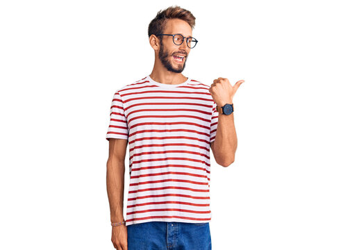 Handsome blond man with beard wearing casual clothes and glasses smiling with happy face looking and pointing to the side with thumb up.