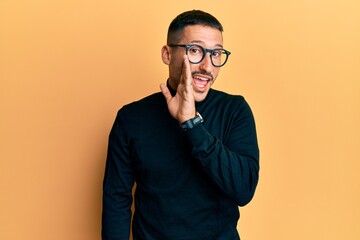 Handsome man with tattoos wearing turtleneck sweater and glasses hand on mouth telling secret rumor, whispering malicious talk conversation