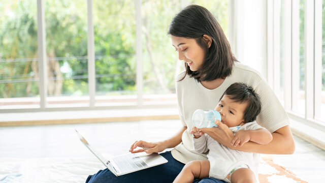 Close Up Of Young Asian Mother On Maternity Leave Trying To Freelance By The Desk With Toddler Child.Stay At Home Mom Working Remotely On Laptop While Taking Care Of Her Baby.