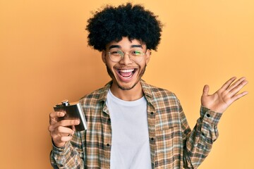 Young african american man with afro hair drinking alcohol from whiskey flask celebrating victory with happy smile and winner expression with raised hands