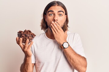 Young handsome man holding chocolate cereals covering mouth with hand, shocked and afraid for...
