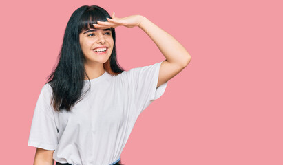 Young hispanic girl wearing casual clothes very happy and smiling looking far away with hand over...