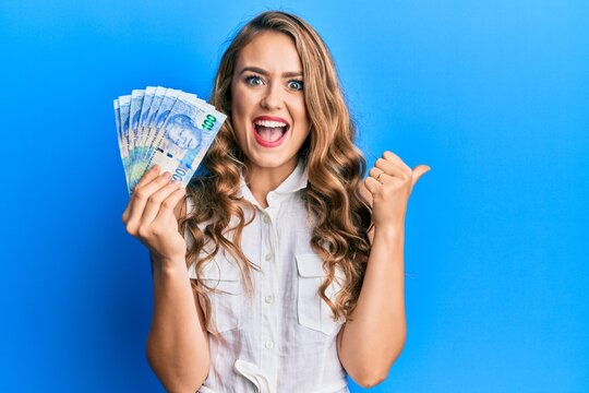 Young blonde girl holding south african 20 rand banknotes pointing thumb up to the side smiling happy with open mouth