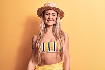 Young beautiful blonde woman wearing bikini and summer hat over isolated yellow background with a happy and cool smile on face. Lucky person.