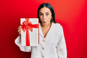Fototapeta premium Young brunette woman holding gift scared and amazed with open mouth for surprise, disbelief face