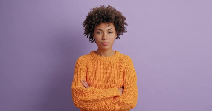 Serious calm ethnic woman with curly hair keeps arms folded winks eyes and looks directly at camera wears casual orange sweater poses against vivid purple background. Bossy person not smiling