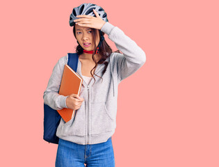 Young beautiful chinese girl wearing student backpack and bike helmet holding book stressed and frustrated with hand on head, surprised and angry face
