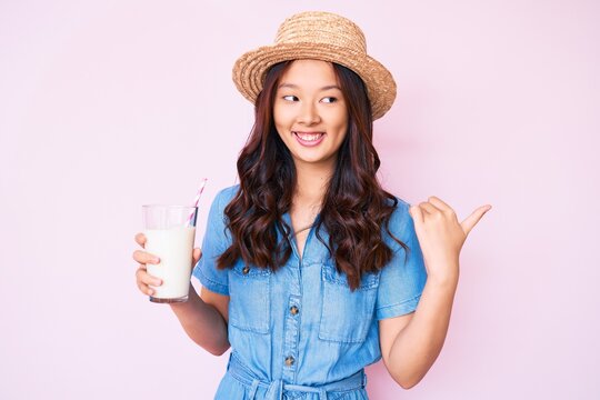 Young beautiful chinese girl wearing summer hat holding glass of milk pointing thumb up to the side smiling happy with open mouth