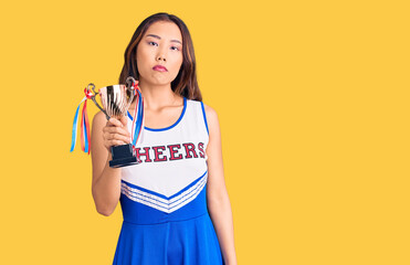 Youn beautiful asian girl wearing cheerleader uniform holding champion trophy thinking attitude and sober expression looking self confident