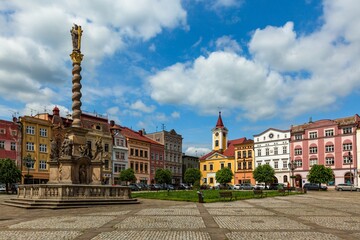 Broumov, Czech Republic - June 17 2020: View of the town square with town hall, colourful houses, green lawn, trees and the Marian column with statues. Sunny summer day with blue sky and clouds.