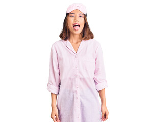 Young beautiful chinese girl wearing sleep mask and pajama sticking tongue out happy with funny expression. emotion concept.