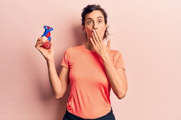 Young beautiful hispanic woman wearing sportswear holding heart covering mouth with hand, shocked...