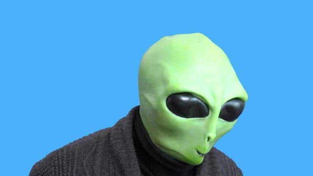 Man in alien costume, warm knitted jacket, grabs his head with his hands, writhes with laughter, against a blue chroma key. Preparation for Halloween party.