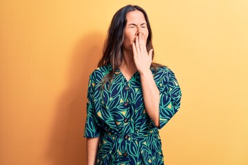 Young beautiful brunette woman wearing casual floral dress standing over yellow background bored yawning tired covering mouth with hand. Restless and sleepiness.