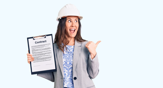 Young beautiful brunette woman wearing architect hardhat holding contract pointing thumb up to the side smiling happy with open mouth