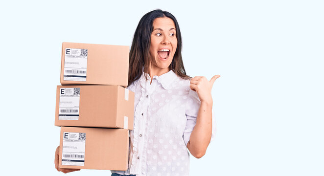 Young beautiful brunette woman holding delivery cardboard package pointing thumb up to the side smiling happy with open mouth