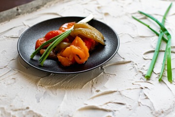 Pickled sweet bell pepper, cut into slices, multicolored vegetables in oil on a black matte plate and a stone white background. The dish is decorated with green leeks