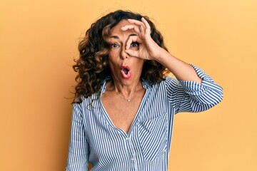 Middle age hispanic woman wearing casual clothes doing ok gesture shocked with surprised face, eye looking through fingers. unbelieving expression.