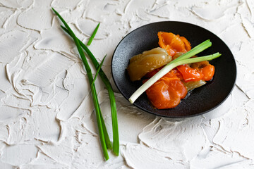 Pickled sweet bell pepper, cut into slices, multicolored vegetables in oil on a black matte plate and a stone white background. The dish is decorated with green leeks