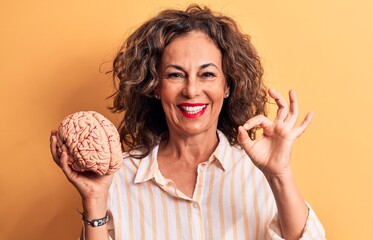Middle age brunette woman asking for care memory holding brain over yellow background doing ok sign...