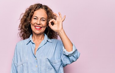Middle age beautiful woman wearing casual denim shirt standing over pink background smiling positive doing ok sign with hand and fingers. Successful expression.