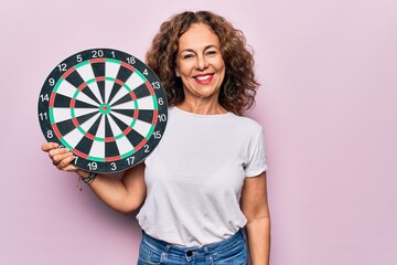 Middle age beautiful woman holding dartboard standing over isolated pink background looking...