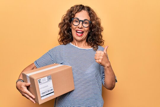 Middle age beautiful woman holding cardboard package with label over yellow background pointing thumb up to the side smiling happy with open mouth