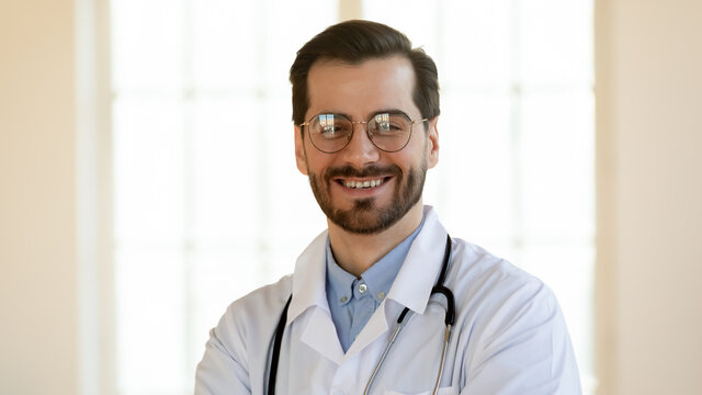 Wide banner panoramic view close up portrait of smiling young Caucasian male doctor in white medical uniform and glasses. Happy man GP or physician in spectacles pose in hospital or clinic.