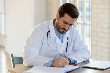 Fototapeta premium Young Caucasian male doctor in white medical uniform sit at desk in hospital write fill patient form in notebook. Focused man GP or physician make notes in clinic medical journal. Healthcare concept.
