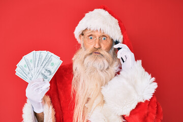 Old senior man wearing santa claus costume holding dollars and smartphone puffing cheeks with funny face. mouth inflated with air, catching air.