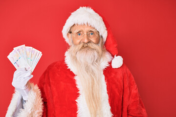 Old senior man with grey hair and long beard wearing santa claus costume holding colombian pesos looking positive and happy standing and smiling with a confident smile showing teeth