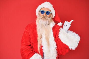 Old senior man wearing santa claus costume and sunglasses smiling happy pointing with hand and finger to the side