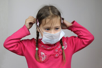 a small blonde girl in a red blouse puts on a medical mask. Gray background