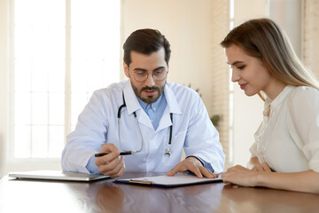 Young male doctor in white medical uniform sit at desk in clinic with female patient discuss illness anamnesis together. Man GP or physician talk consult woman client, consider results or treatment.