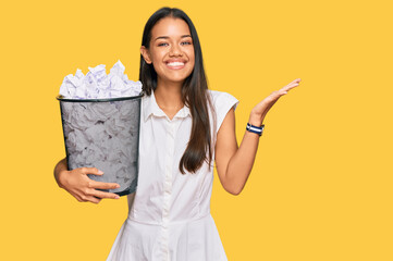 Beautiful hispanic woman holding paper bin full of crumpled papers celebrating victory with happy...