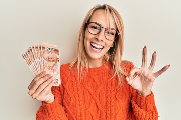 Beautiful blonde woman holding 50 turkish lira banknotes doing ok sign with fingers, smiling...