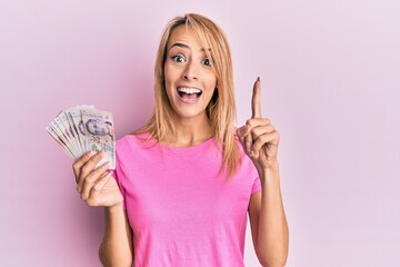 Beautiful blonde woman holding 2 singapore dollars banknotes smiling with an idea or question pointing finger with happy face, number one