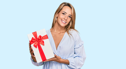Beautiful blonde woman holding gift looking positive and happy standing and smiling with a confident smile showing teeth