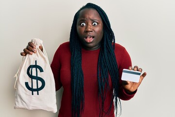 Young black woman with braids holding dollars bag and credit card afraid and shocked with surprise...