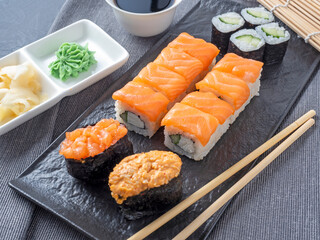 a variety of Japanese rolls and sushi on a textured plate. Side view. bamboo sticks of ginger and sauce nearby.