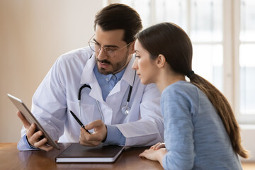 Young Caucasian male doctor and female patient look at tablet screen discuss test results on gadget together. Man GP or physician use pad explain anamnesis to woman client at consultation in hospital. - obrazy, fototapety, plakaty