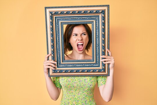 Young beautiful woman holding empty frame angry and mad screaming frustrated and furious, shouting with anger. rage and aggressive concept.