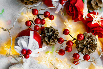 Christmas decorations on a soft cozy white blanket. Red toys in the form of balls and berries, with fir cones. Around yellow lanterns light bulbs in the form of stars. Elegant composition for Christma