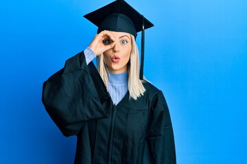 Beautiful blonde woman wearing graduation cap and ceremony robe doing ok gesture shocked with surprised face, eye looking through fingers. unbelieving expression.