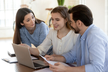 Smiling diverse employees sit at desk in office work on laptop discussing paperwork together. Happy colleagues coworkers brainstorm cooperate on computer chatting talking. Teamwork concept.