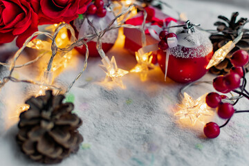 Christmas decorations on a soft cozy white blanket. Red toys in the form of balls and berries, with fir cones. Around yellow lanterns light bulbs in the form of stars. Elegant composition for Christma