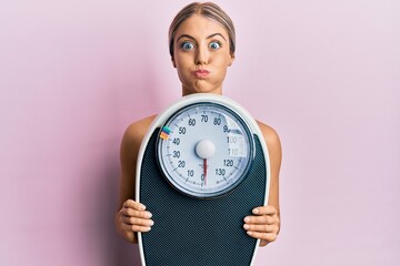 Beautiful blonde woman holding weight machine to balance weight loss puffing cheeks with funny...